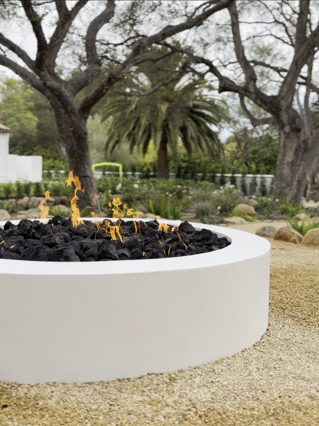 Round white concrete fire pit with lava rock alight, under oaks on a decomposed-granite terrace