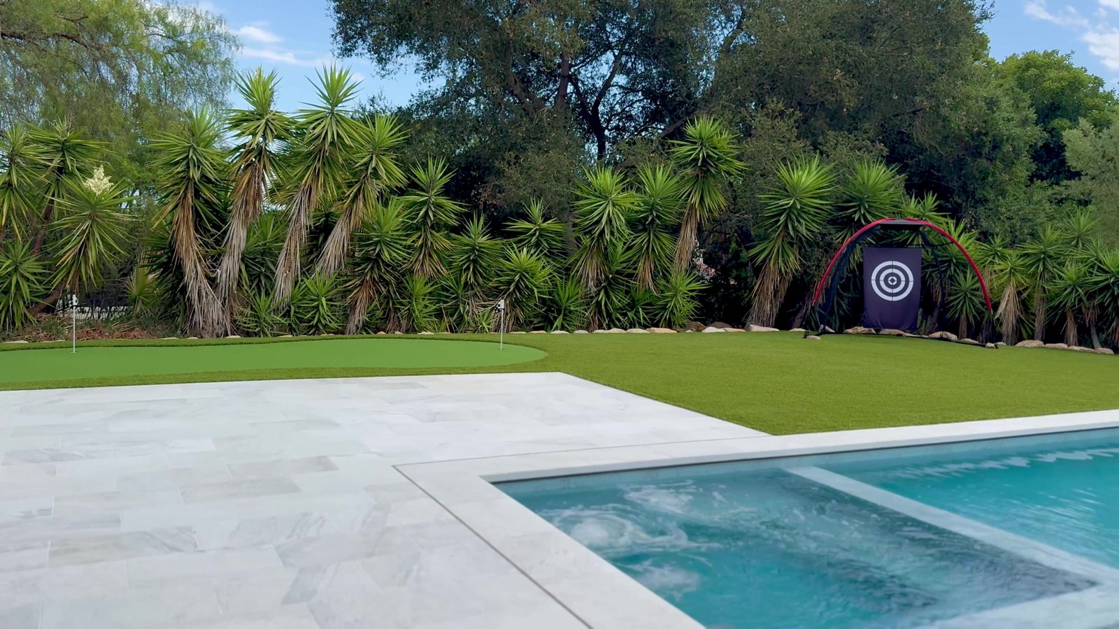 Pool and spa with white paving beside a putting green and palms